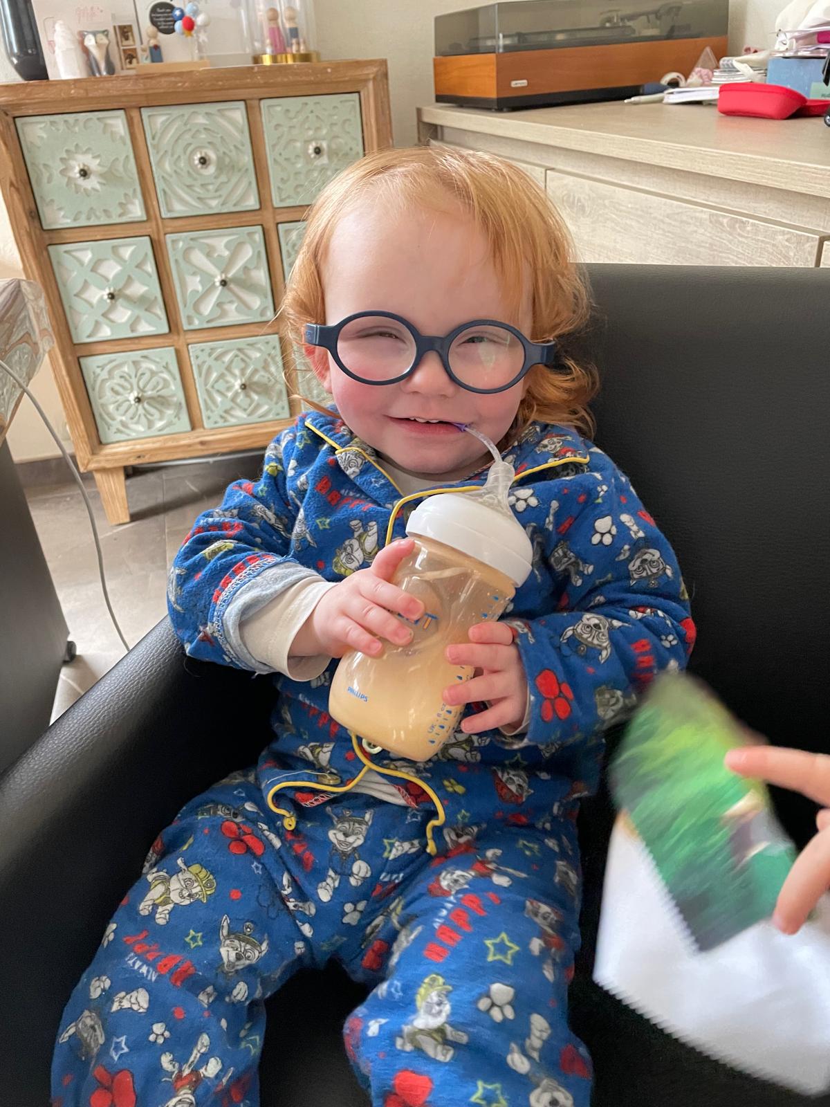 Seth in pyjama met bril lacht terwijl hij zijn melkfles vasthoudt